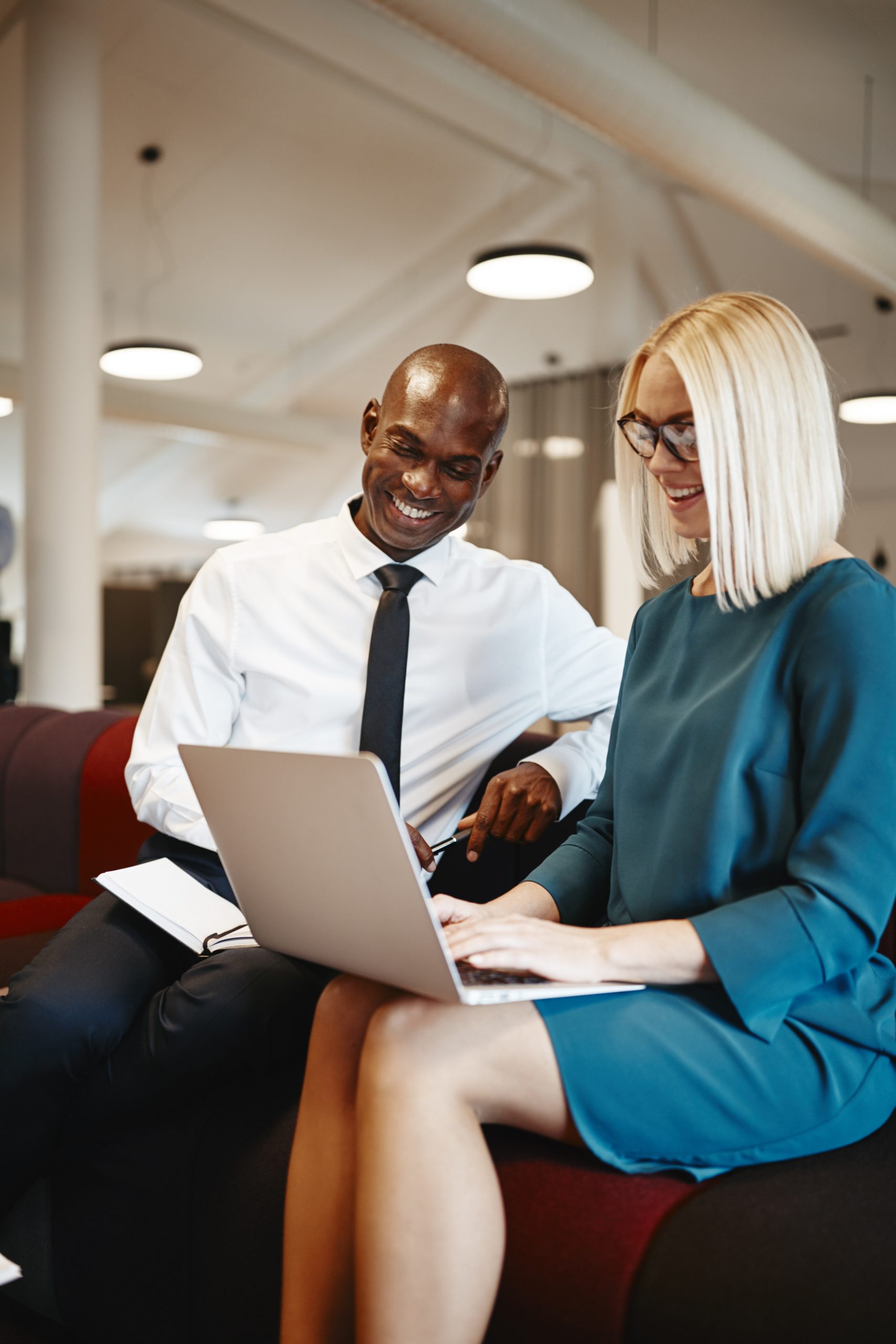 Two diverse business colleagues smiling while sitting together on a sofa in a modern office working online with a laptop